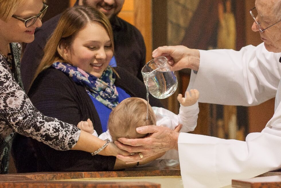 A priest pours water over a babys head in a baptism ceremony as adults hold the baby and watch attentively.