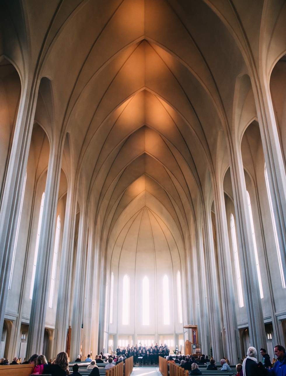Large modern church interior with high vaulted ceilings, tall arched windows, and people seated in pews facing the altar.