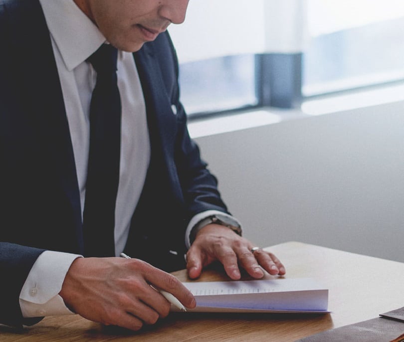 A person in a suit sits at a desk by a window with natural light, turning the page of a document or book.