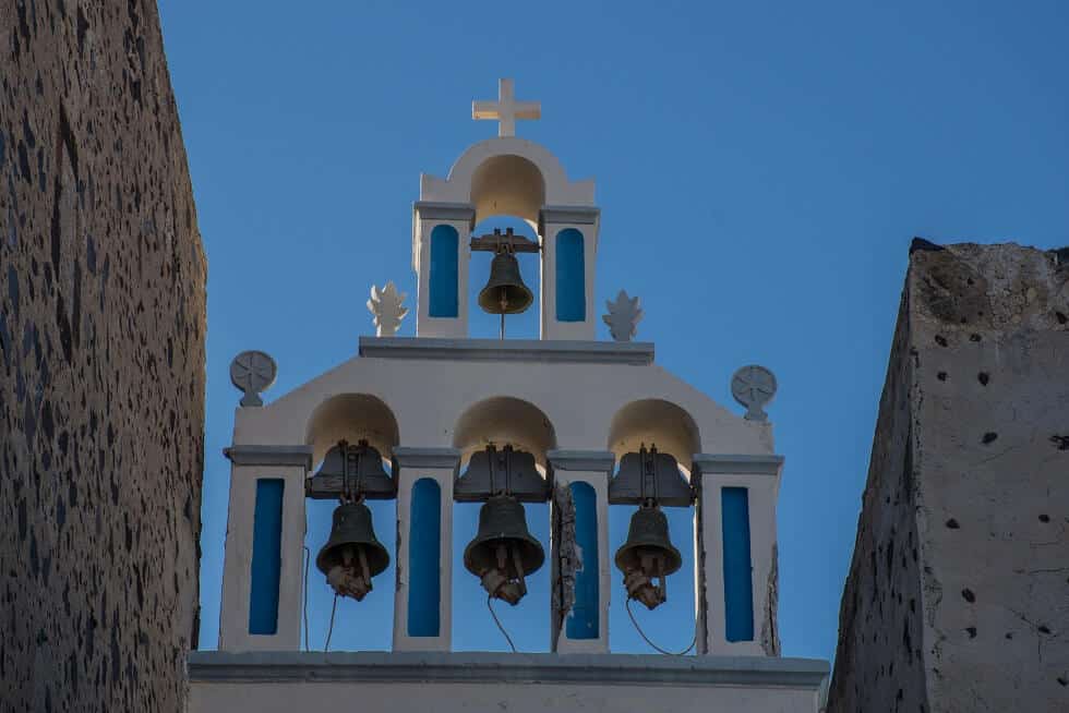 White church bell tower with blue accents and a cross, positioned between two stone walls under a clear blue sky.