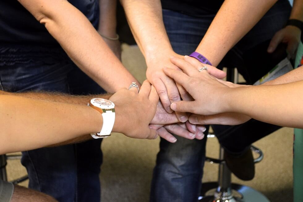 Six people seated on stools stack their hands together, showing teamwork. Some wear watches or rings.