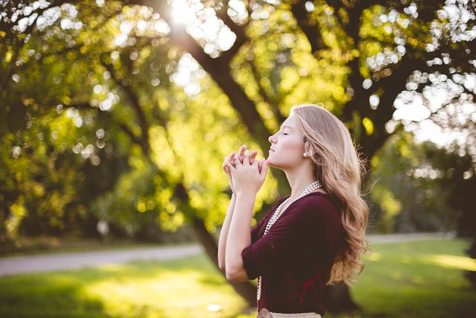A woman with long hair stands in a sunlit park, eyes closed, hands clasped in front of her as if praying or contemplating.