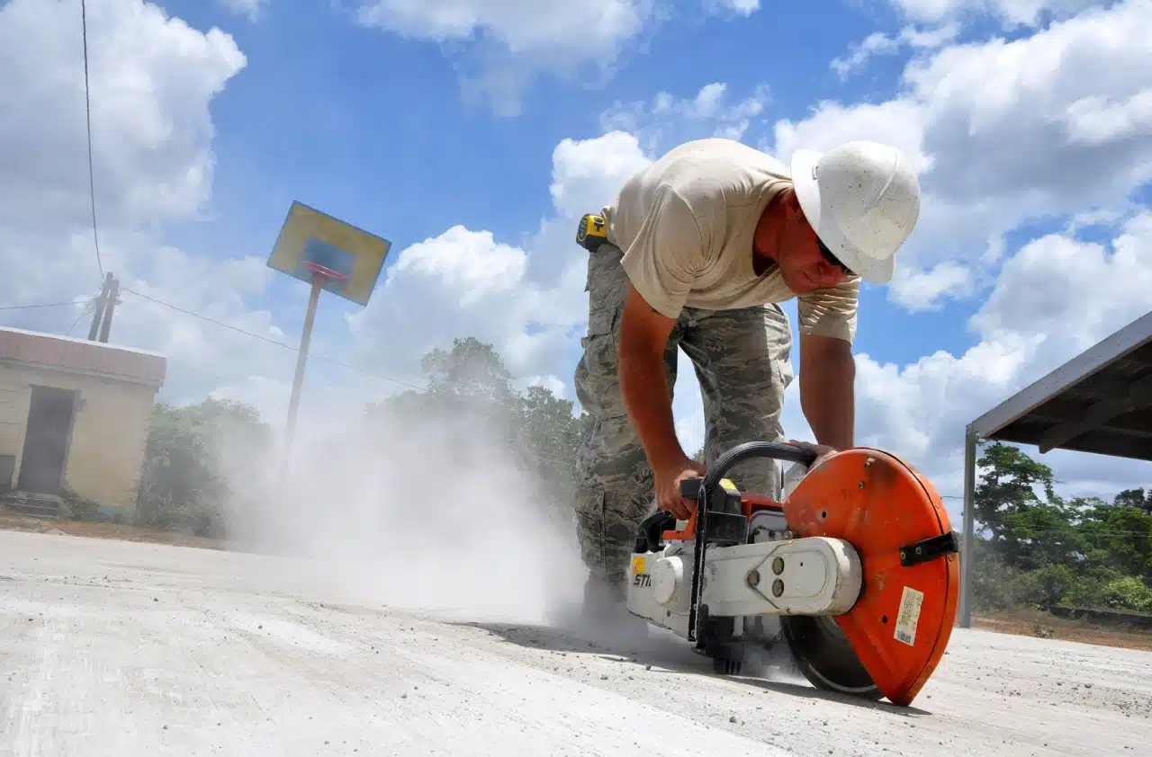 Construction worker in hard hat uses a power saw to cut outdoor concrete, making dust under a blue sky with scattered clouds.