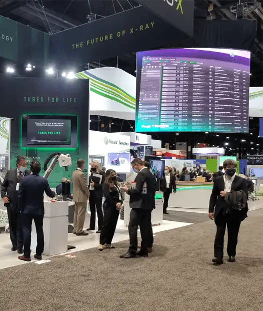 Attendees browse booths and digital displays at a tech trade show, with a large screen showing data and signs about X-ray technology.