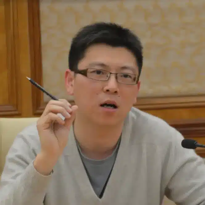 Person with glasses and a light sweater holds a pen while speaking at a meeting, seated beside a microphone.
