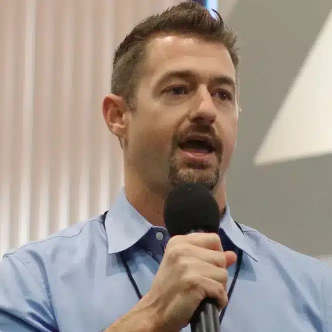 Man in a blue collared shirt speaks into a microphone indoors; background is blurred.