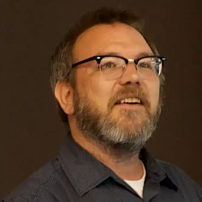 Man with glasses and beard, in a dark collared shirt, looks slightly upward and smiles against a dark background.