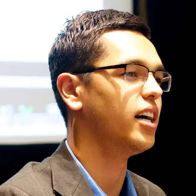 A man with short dark hair and glasses, in a suit jacket and blue shirt, speaks before a blurred screen.