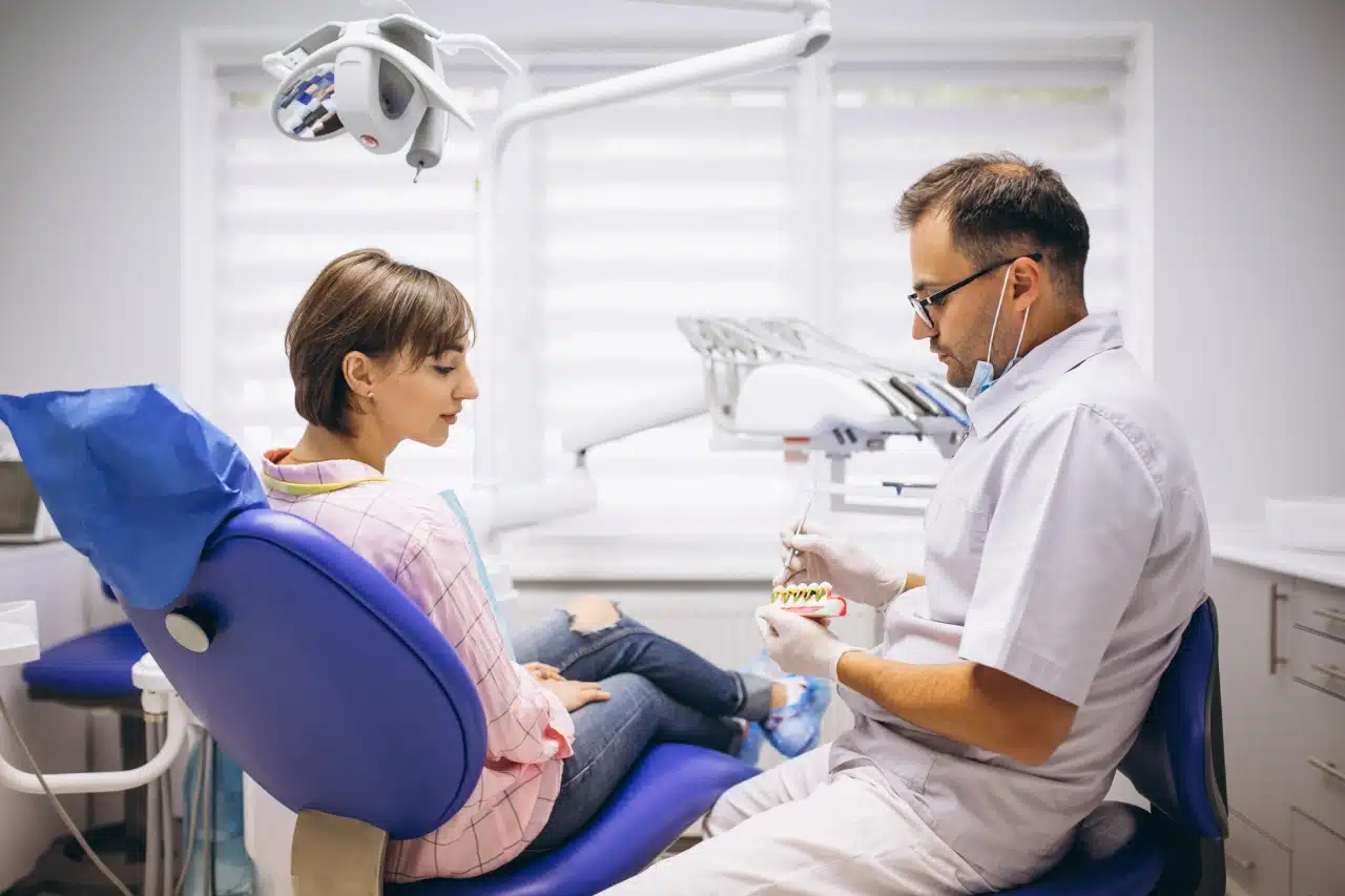 Dentist in white coat demonstrates a dental model to a seated female patient in a modern dental clinic.