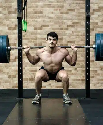 Man in shorts and sneakers performs a back squat with a loaded barbell in a gym, brick wall visible in the background.