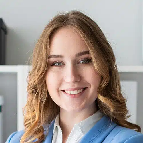 Young woman with wavy light brown hair, smiling at the camera in a light blue blazer and white shirt, standing indoors by shelves.