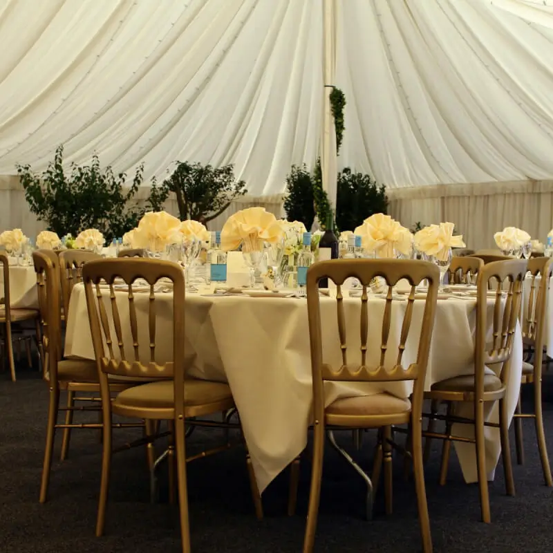Round tables with cream tablecloths and folded napkins sit under a white tent canopy, with chairs and greenery in the background.