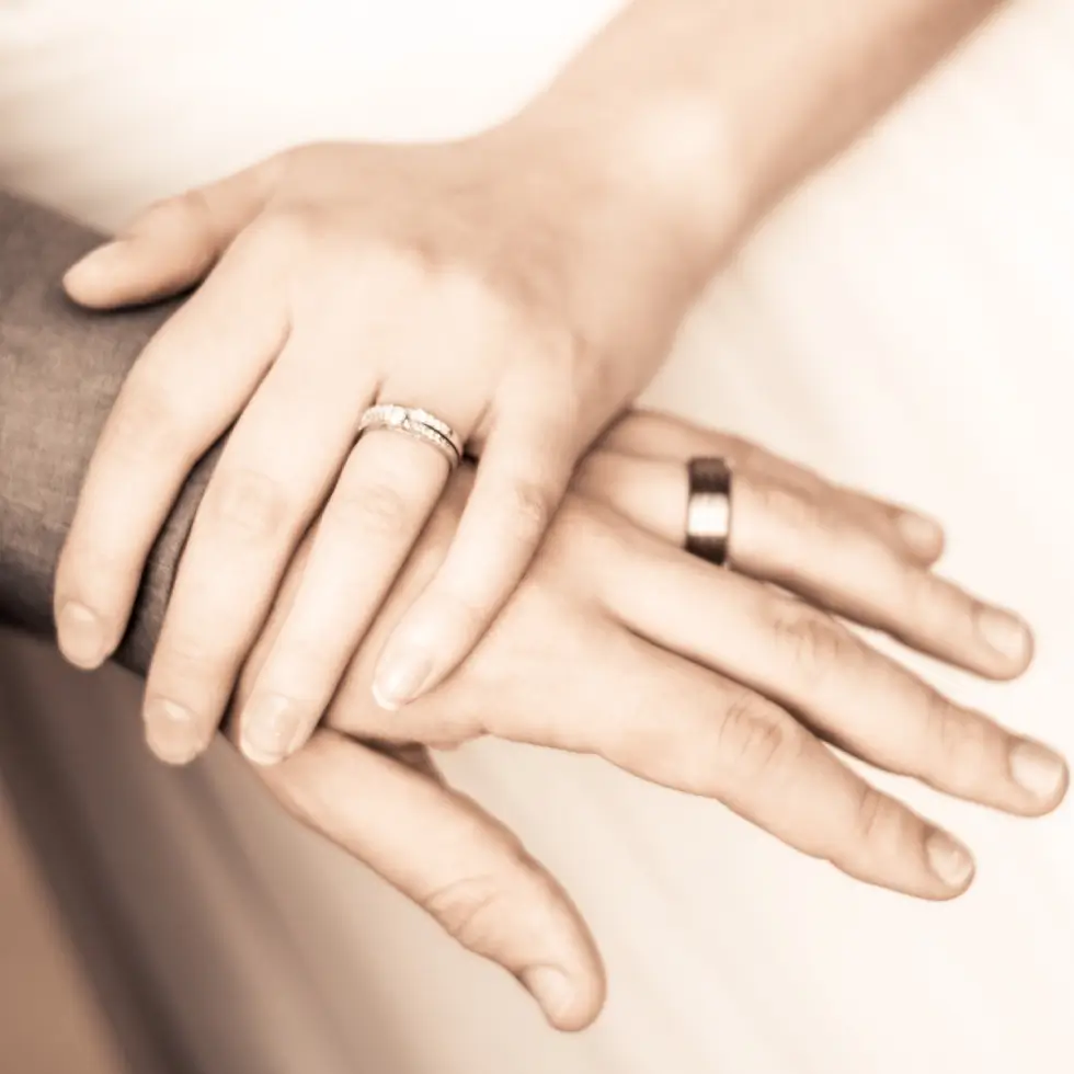 Two hands with wedding rings rest on each other—one hand has a silver ring, the other a dark metallic band.