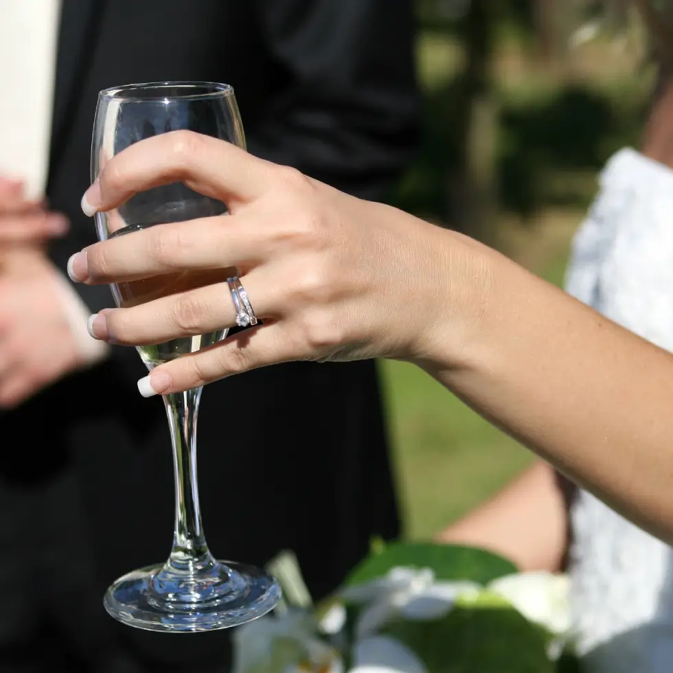 A person with a diamond ring holds a champagne glass; another person in formal attire stands blurred in the background.
