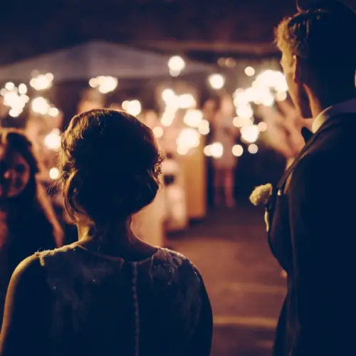 Bride and groom stand together facing guests holding sparklers at an outdoor nighttime wedding celebration.