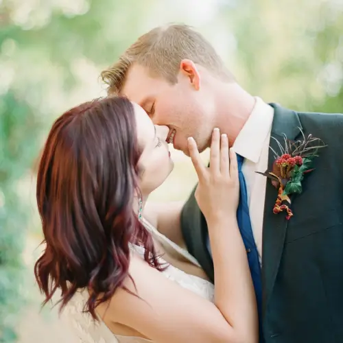 A couple in wedding attire embraces outdoors, gazing at each other closely. The groom wears a suit; the bride touches his face.