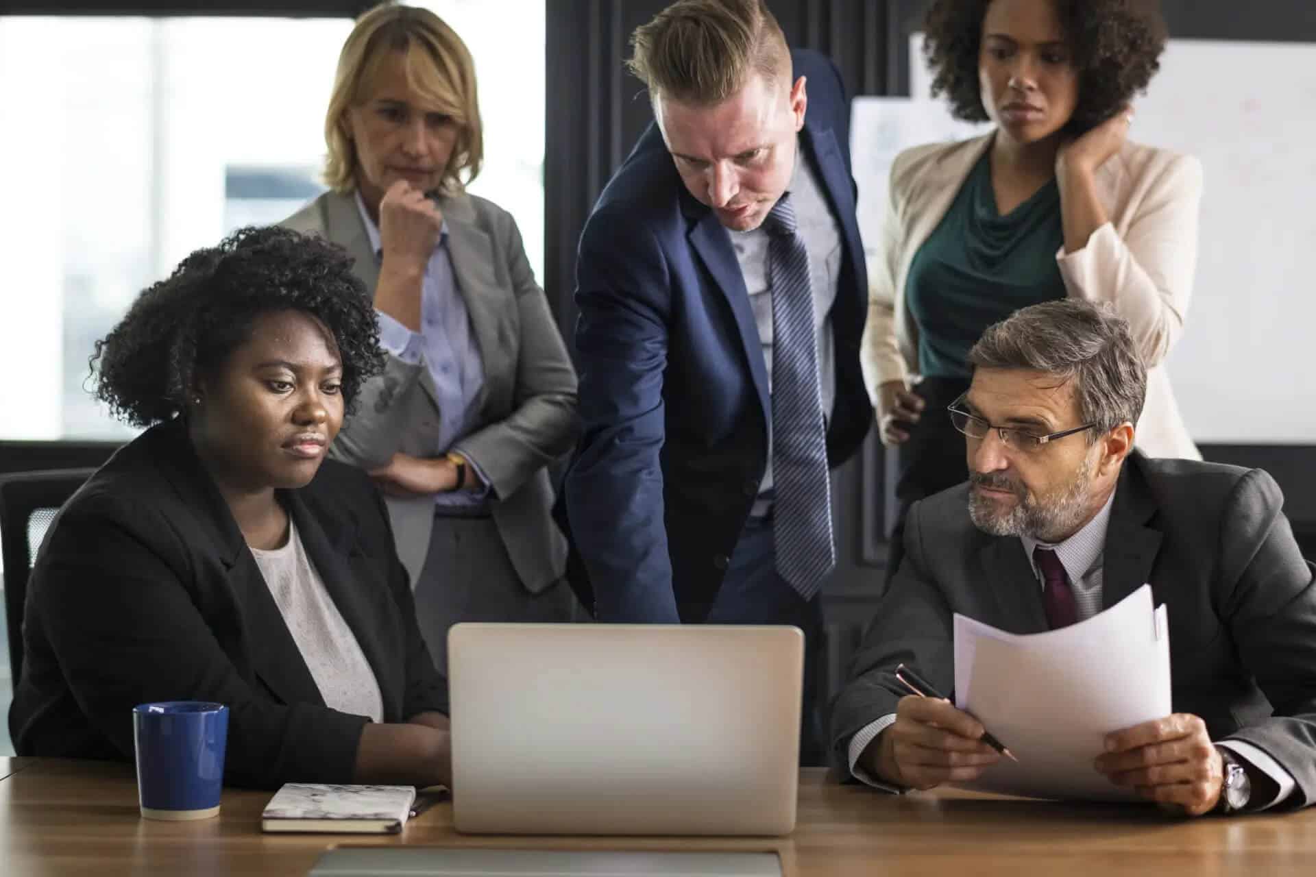 Five professionals in business attire gather around a laptop in an office, discussing or reviewing the screens content together.