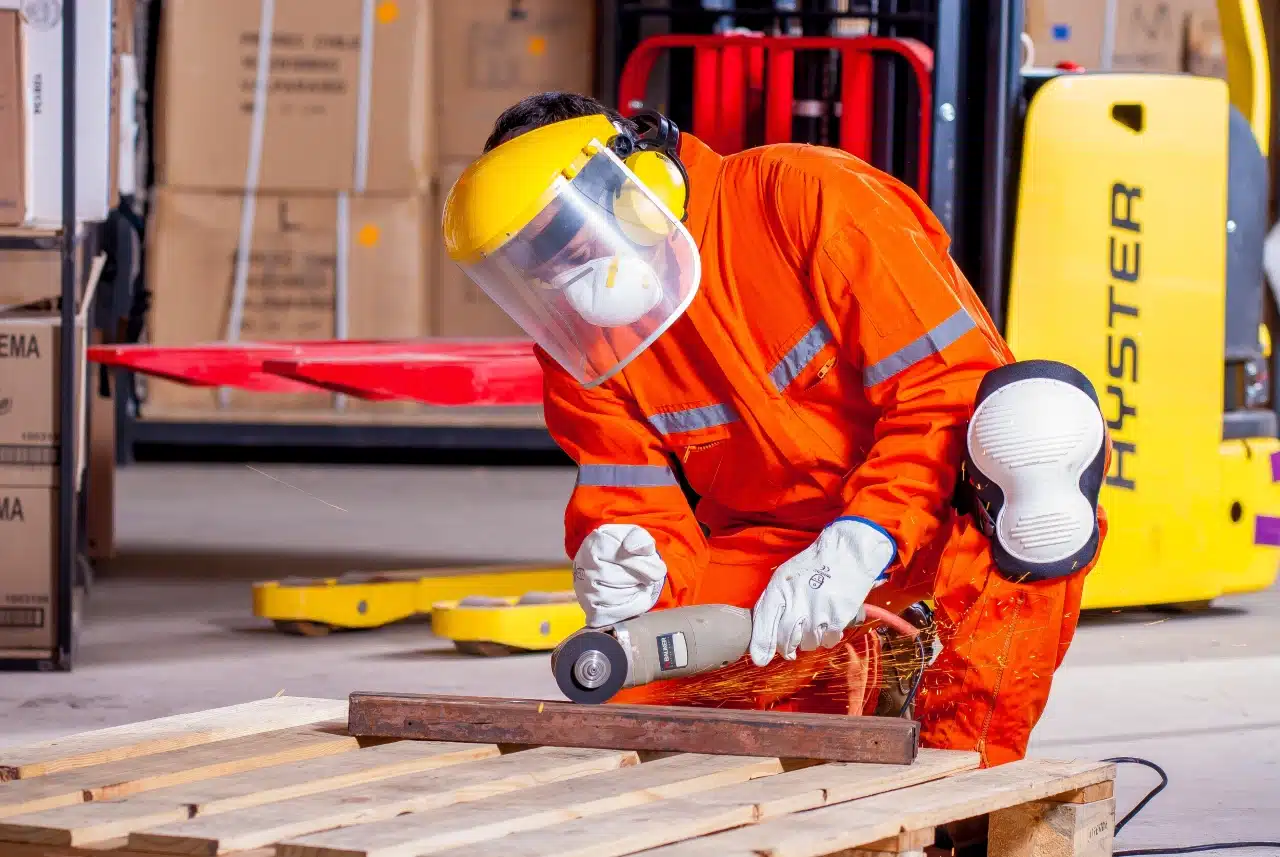 Worker in orange safety gear and face shield grinding metal on a wooden pallet with a power tool in a warehouse.