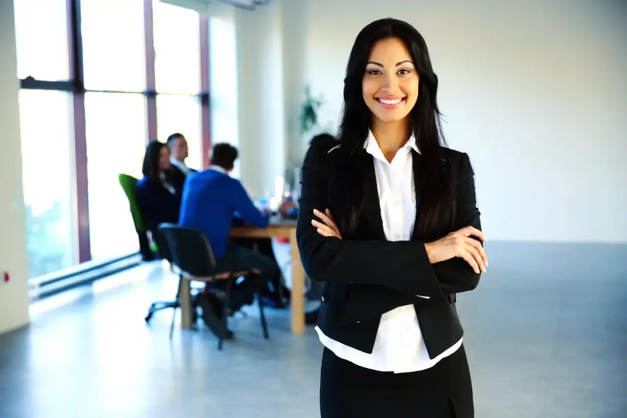 Smiling woman in business attire stands with arms crossed in front of an office, people working at a table in the background.