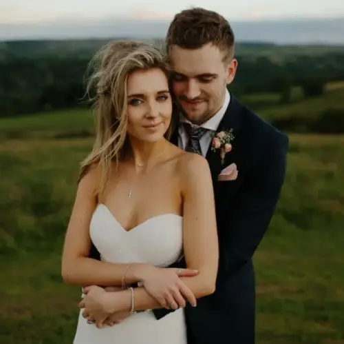 Bride in strapless white dress and groom in dark suit embrace outdoors, with green fields visible behind them.
